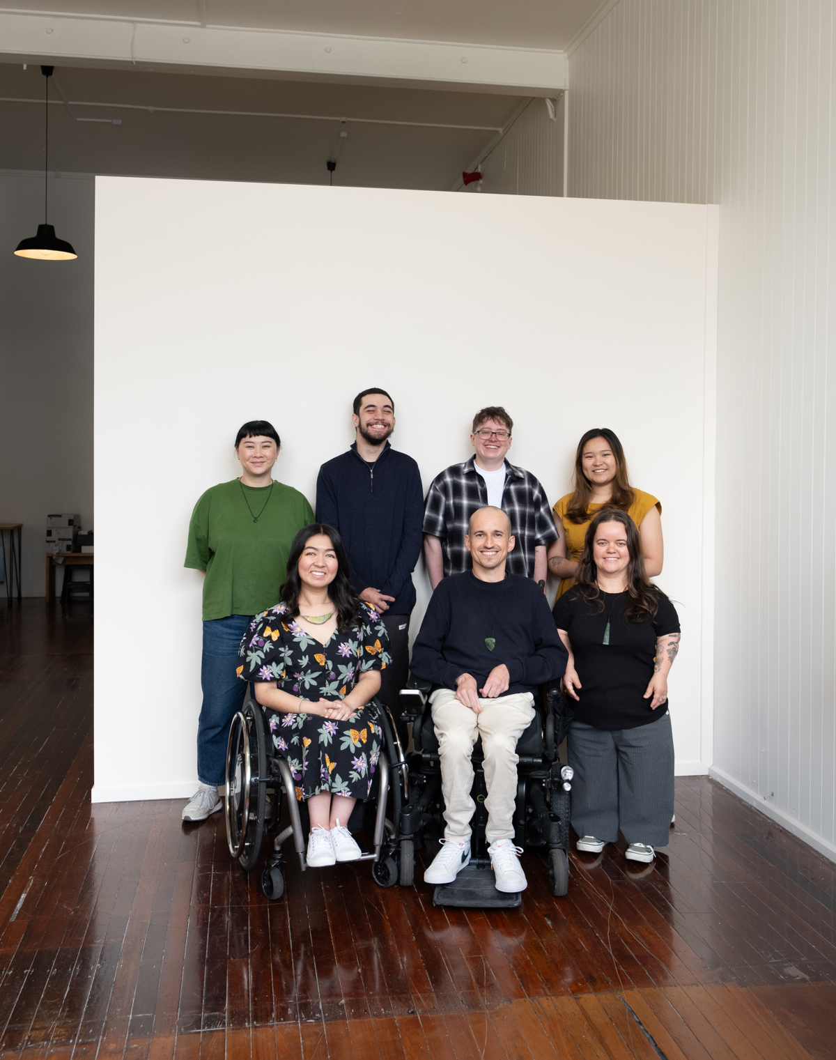 The D*List team sit and stand for a group photo on a wooden floor and against a white wall.